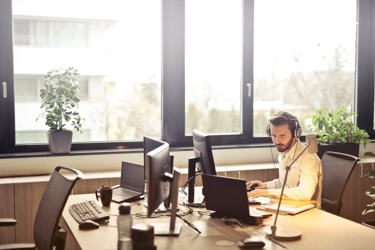 Ein Mann mit Kopfhörern sitzt an einem Schreibtisch mit mehreren Computermonitoren und einem Laptop und nutzt eine Kundenbeziehungsmanagement-Software in einem modernen Büro mit großen Fenstern und Topfpflanzen.