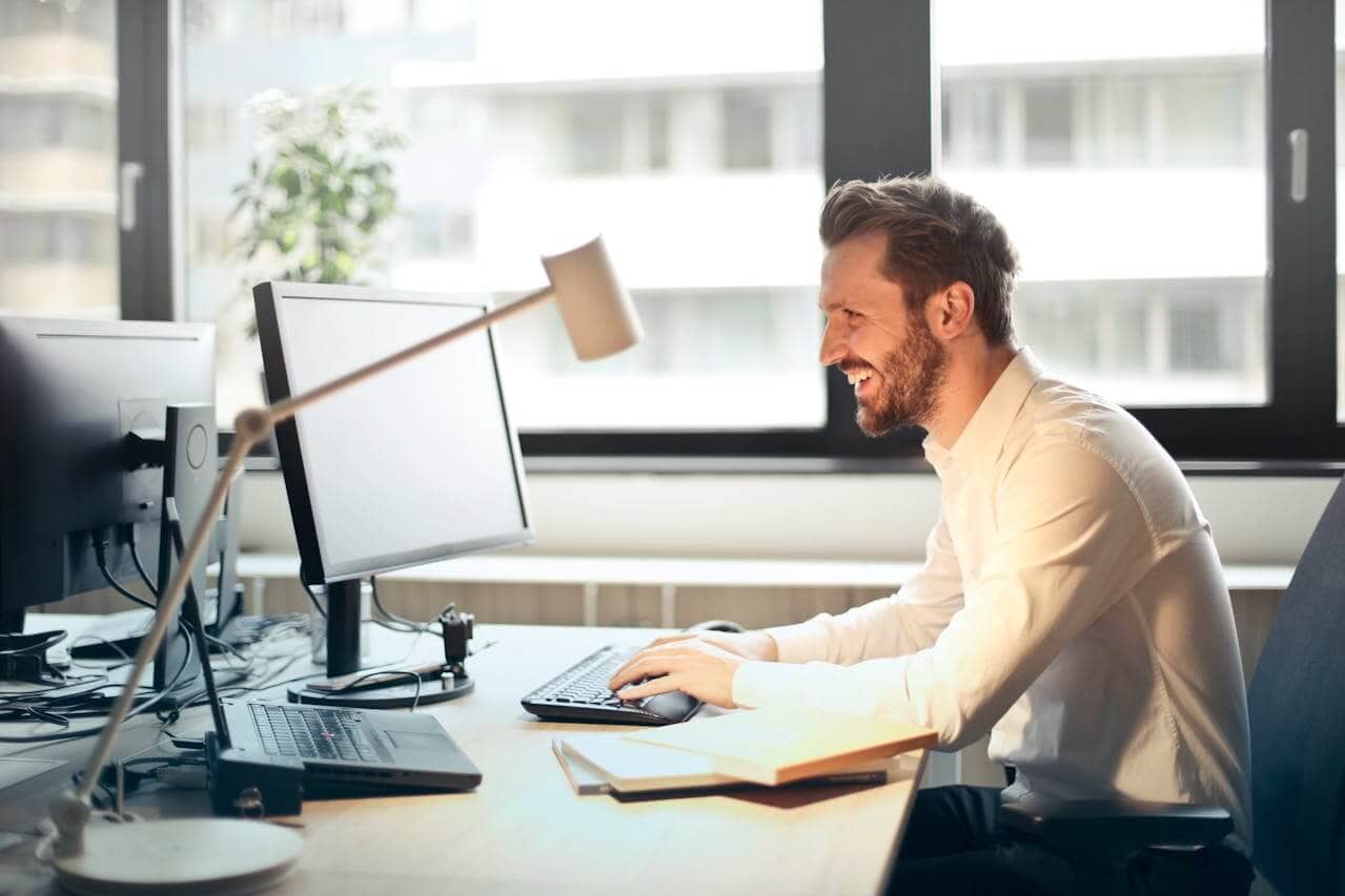 Ein Mann im weißen Hemd sitzt lächelnd an einem Schreibtisch und tippt auf einer Tastatur. Er befindet sich in einem modernen Büro mit großen Fenstern, zwei Monitoren, auf denen E-Mail-Marketing-Dashboards angezeigt werden, einer Lampe und einem Buch auf dem Schreibtisch.