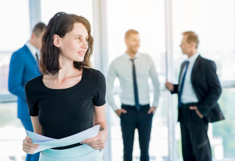 Eine Frau mit Papieren steht selbstbewusst in einem Büro und stellt ihren Ruf als Führungskraft zur Schau. Im Hintergrund sind drei Männer in ein Gespräch vertieft.