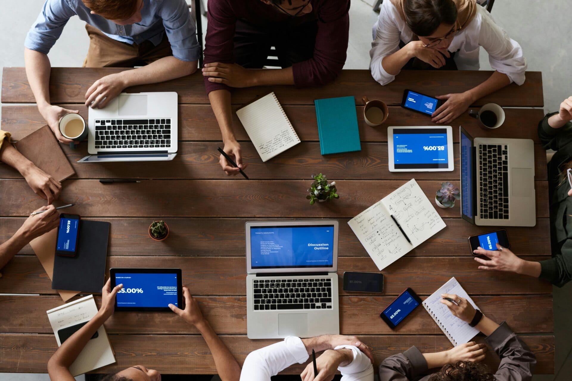 Eine Gruppe von Menschen sitzt mit Laptops, Tablets, Notebooks und Smartphones um einen Holztisch, arbeitet und tauscht Informationen in einer kollaborativen Clownswelt-Umgebung aus.