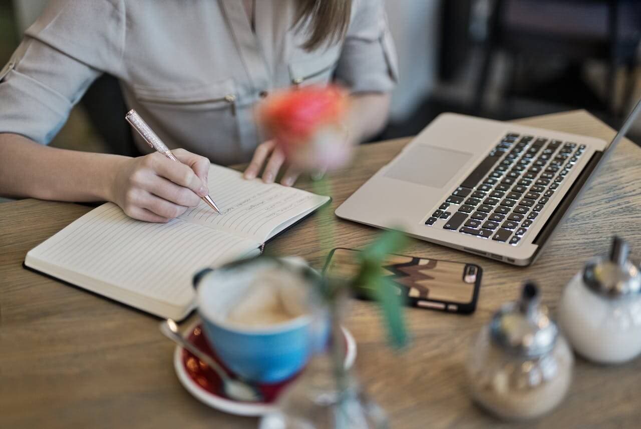 Person, die einzigartige Inhalte in einem Notizbuch erstellt, neben einem offenen Laptop und einer Tasse Kaffee auf dem Tisch.