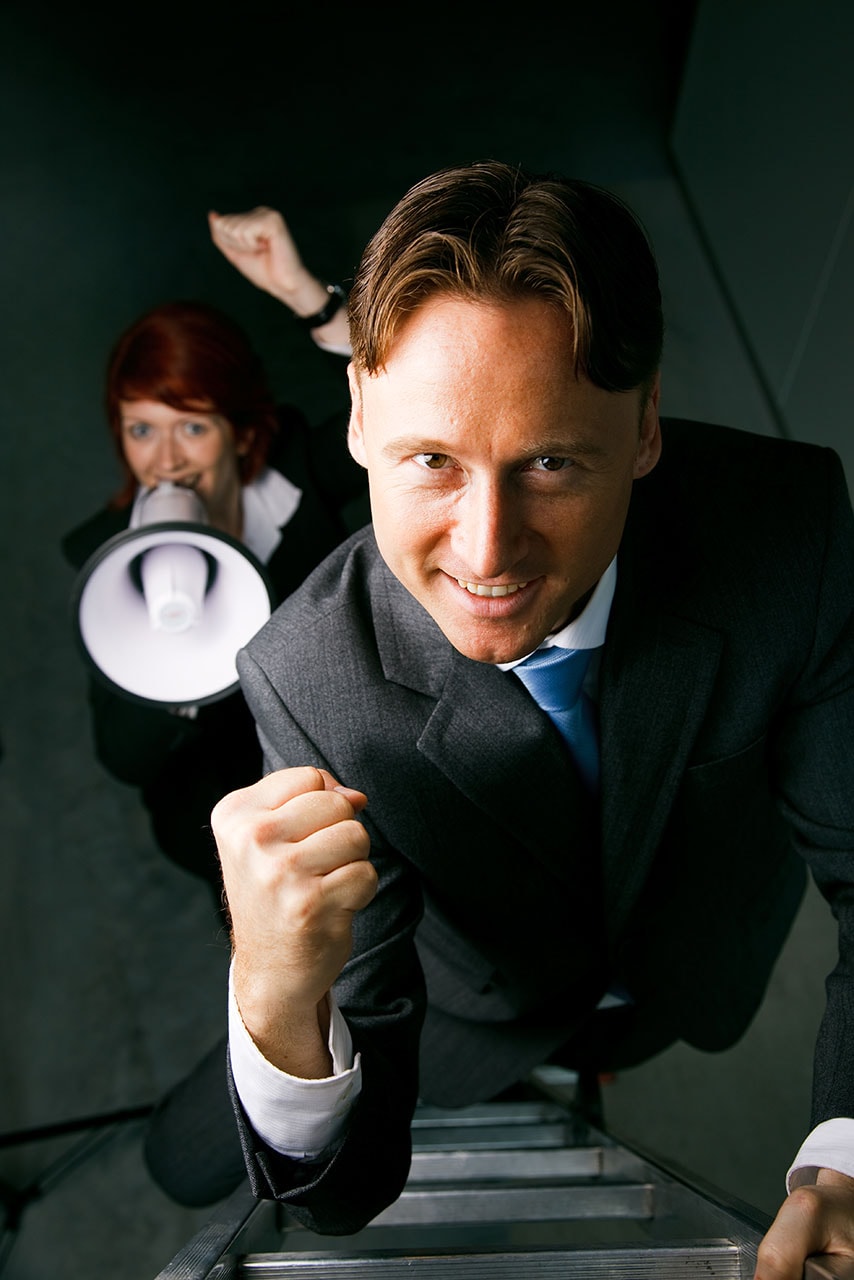 Ein Mann im Anzug klettert auf eine Leiter und macht eine Geste mit der Faust, während eine Frau hinter ihm durch ein Megaphon schreit. Beide blicken nach oben und symbolisieren das Streben nach Erfolg, das man oft in Native Ads-Kampagnen sieht.