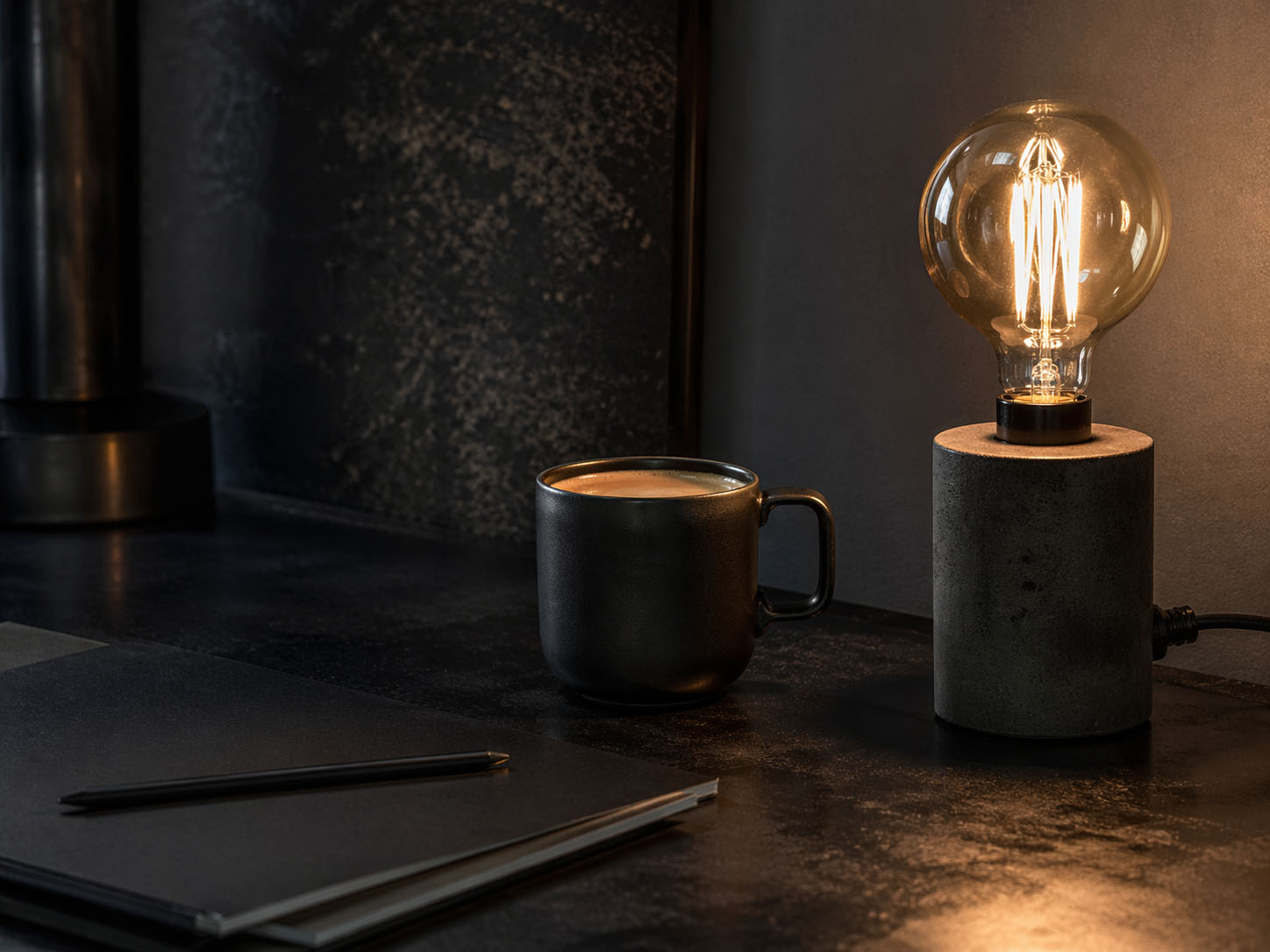 Ein schwarzer Kaffeebecher steht neben einer brennenden Glühbirnenlampe auf einem dunklen Schreibtisch, auf dem ein geschlossenes Notizbuch und ein Stift liegen, perfekt, um Gedanken zu notieren oder unerwünschte Einträge zu vermerken.
