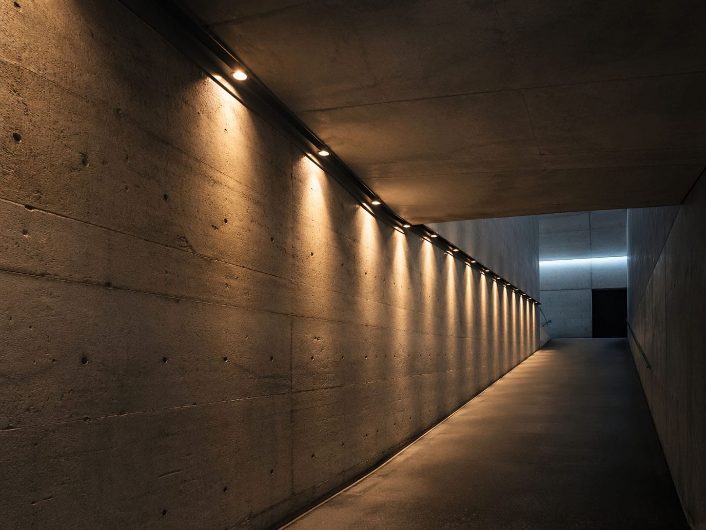 Ein Betonkorridor mit einer Rampe, die von Deckeneinbaustrahlern an der linken Wand beleuchtet wird, führt zu einer geschlossenen dunklen Tür am Ende, die keine Chance für unerwünschte Einträge bietet.