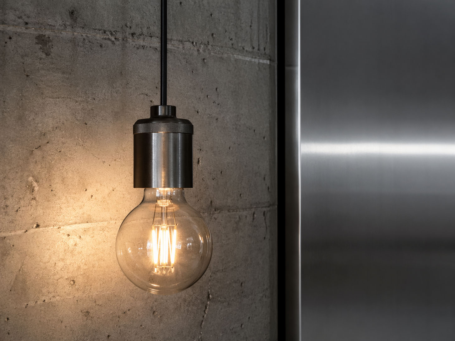 Eine brennende Glühbirne mit Metallfassung hängt vor einer Betonwand neben einer gebürsteten Metalloberfläche, beleuchtet den Bereich und enthüllt sogar unerwünschte Einträge auf dem strukturierten Hintergrund.