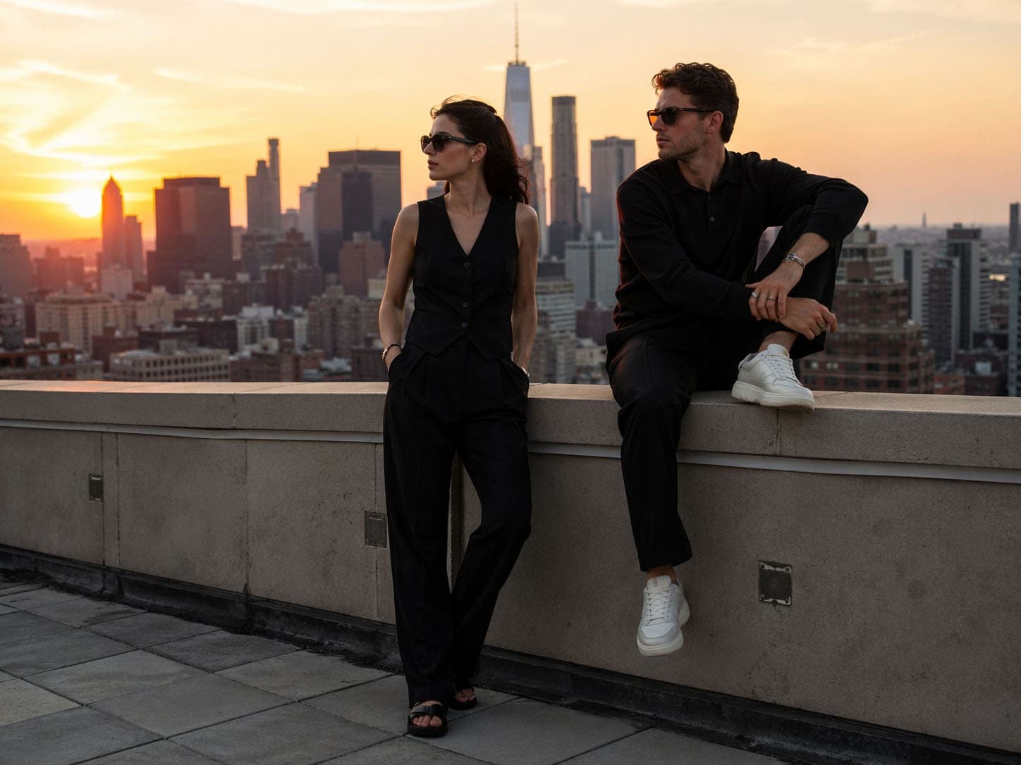 Zwei stilvolle Personen auf einer Dachterrasse mit Blick auf die Skyline bei Sonnenuntergang.