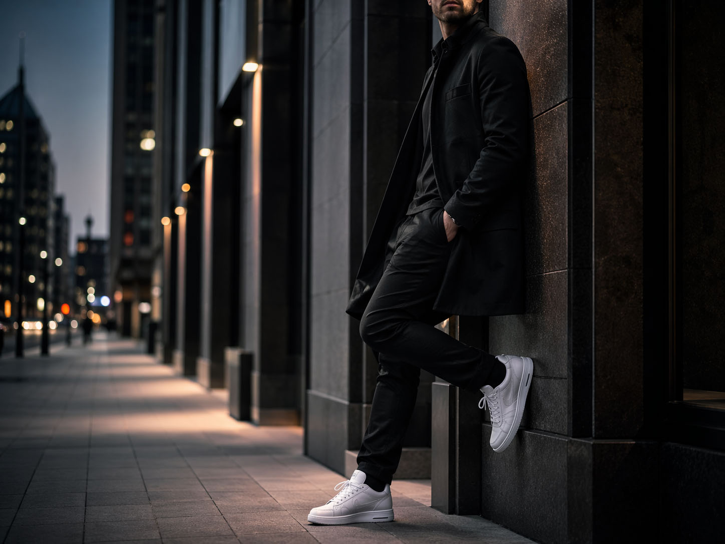 Mann in schwarzem Mantel und weißen Sneakers lehnt an einer Wand in der Stadt bei Nacht.