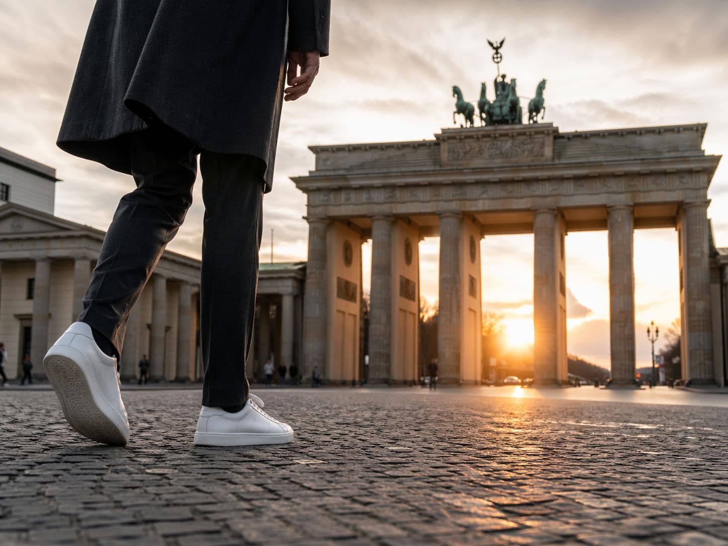 Person, die bei Sonnenuntergang in Berlin auf das Brandenburger Tor zugeht, gesehen aus einem niedrigen Winkel mit Fokus auf ihre weißen Turnschuhe und den Kopfsteinpflasterboden, frei von unerwünschten Einträgen für eine saubere und fesselnde Szene.