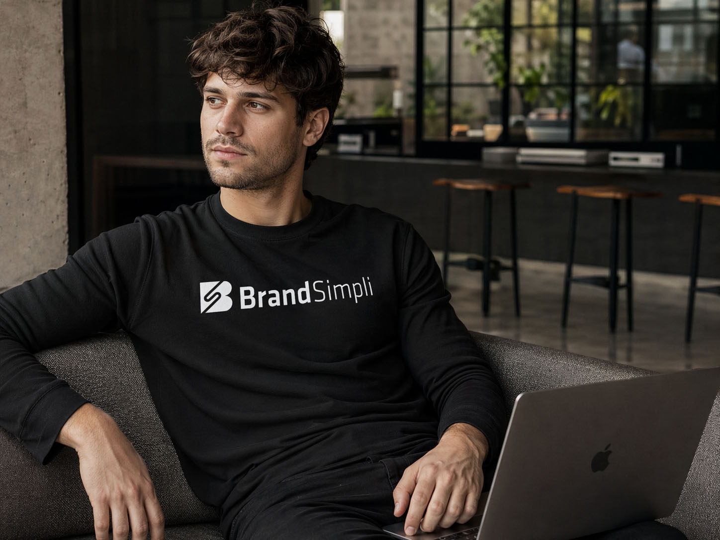 Ein Mann mit zerzaustem Haar sitzt auf einer Couch neben einem Laptop, trägt ein schwarzes "BrandSimpli"-Sweatshirt und schaut in einem modernen Büro zur Seite - konzentriert darauf, unerwünschte Einträge in einem gut beleuchteten Arbeitsbereich zu bearbeiten.