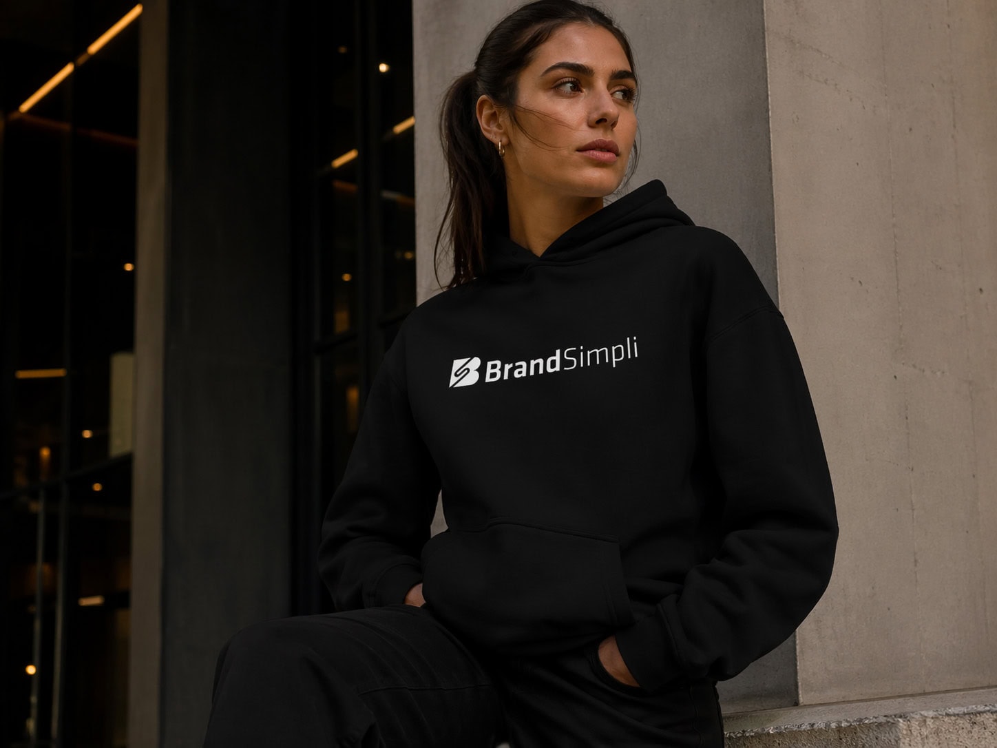 Eine Frau, die einen schwarzen Kapuzenpulli mit dem "BrandSimpli"-Logo und einem LP Analyse-Aufnäher trägt, sitzt draußen an einer Betonwand und schaut zur Seite.