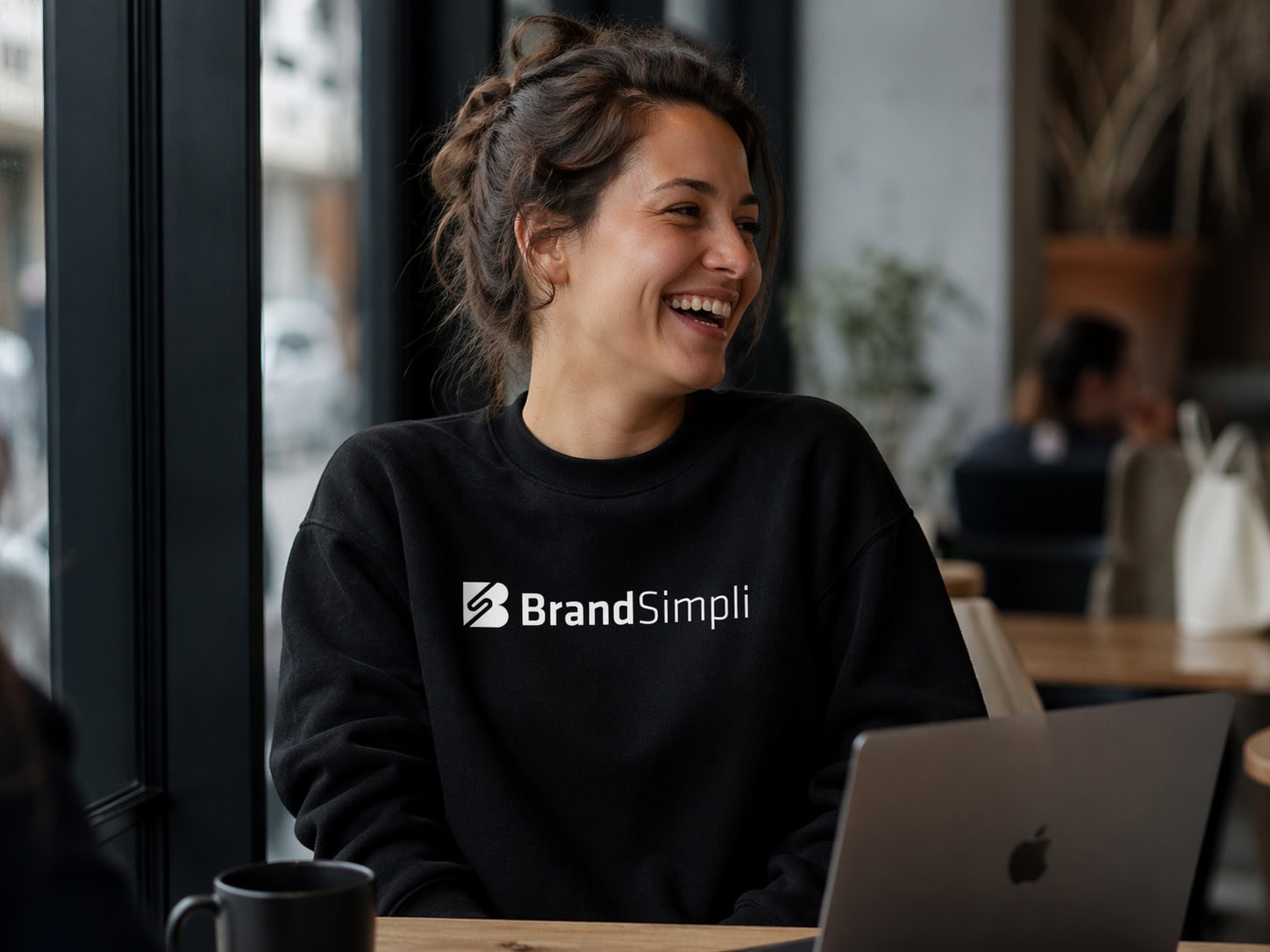 Lächelnde Frau, die mit einem Laptop an einem Tisch sitzt und ein schwarzes "BrandSimpli"-Sweatshirt in einem Café trägt, bereit, digitale PR-Strategien in Angriff zu nehmen.