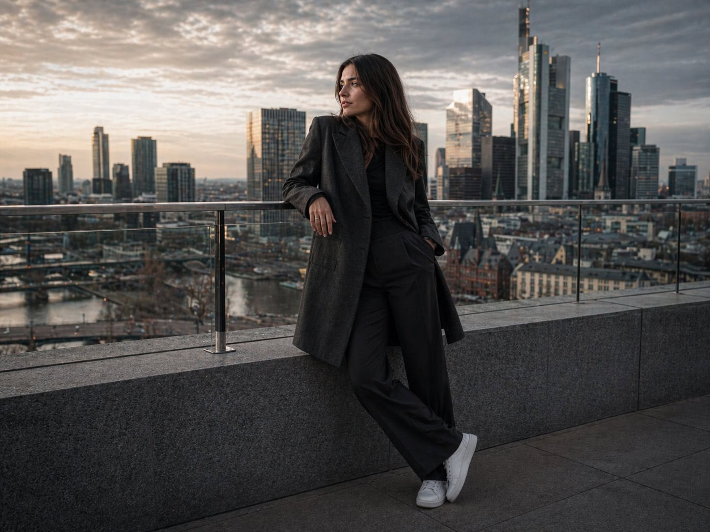 Eine Frau in dunkler Kleidung lehnt sich an einen Betonsims und verkörpert das Selbstvertrauen der digitalen PR, mit einer Stadtsilhouette und hohen Gebäuden im Hintergrund bei Sonnenuntergang.