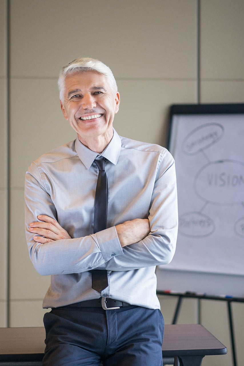 Ein grauhaariger Mann mit Hemd und Krawatte steht mit verschränkten Armen lächelnd vor einem Flipchart, auf dem das Wort VISION", ein einfaches Diagramm und Notizen zu effektiven Native-Ads-Strategien stehen.